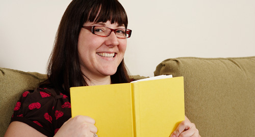 A photo of a woman wearing glasses. She is holding a book and smiling.