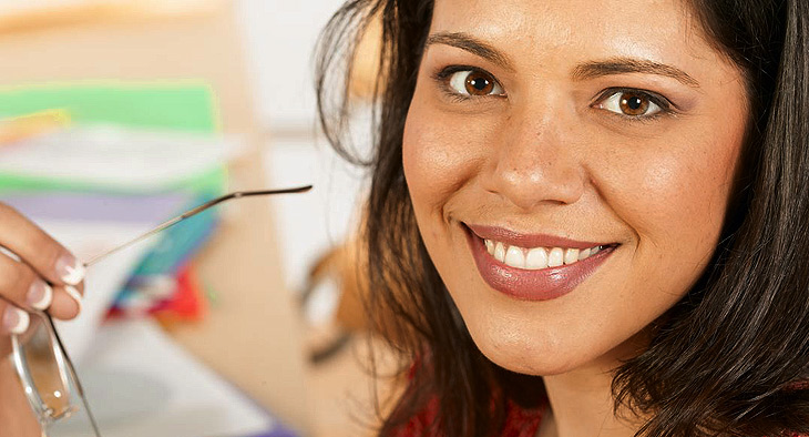 A photo of Tasha, a woman with long dark hair and brown eyes. She is smiling and holding her glasses.