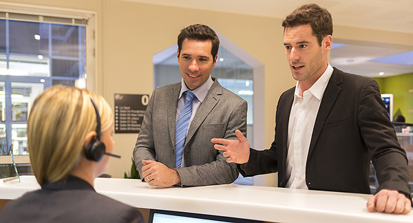 A photo of two people at an information counter. They are asking a woman for information.