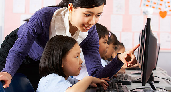 A photo of a teacher in class with young students. The students are sitting in a line at computers. The teacher is pointing to a computer screen amd helping one of the students. 