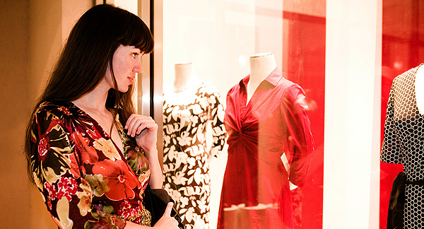 A photo of a woman in a floral dress looking in shop window at dresses on shop dummies.