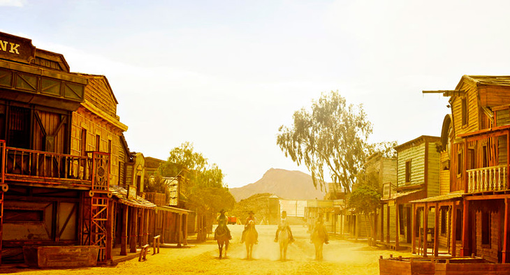A photo of a film set for a western. There is the street of a town and four men on horses riding along the street. 